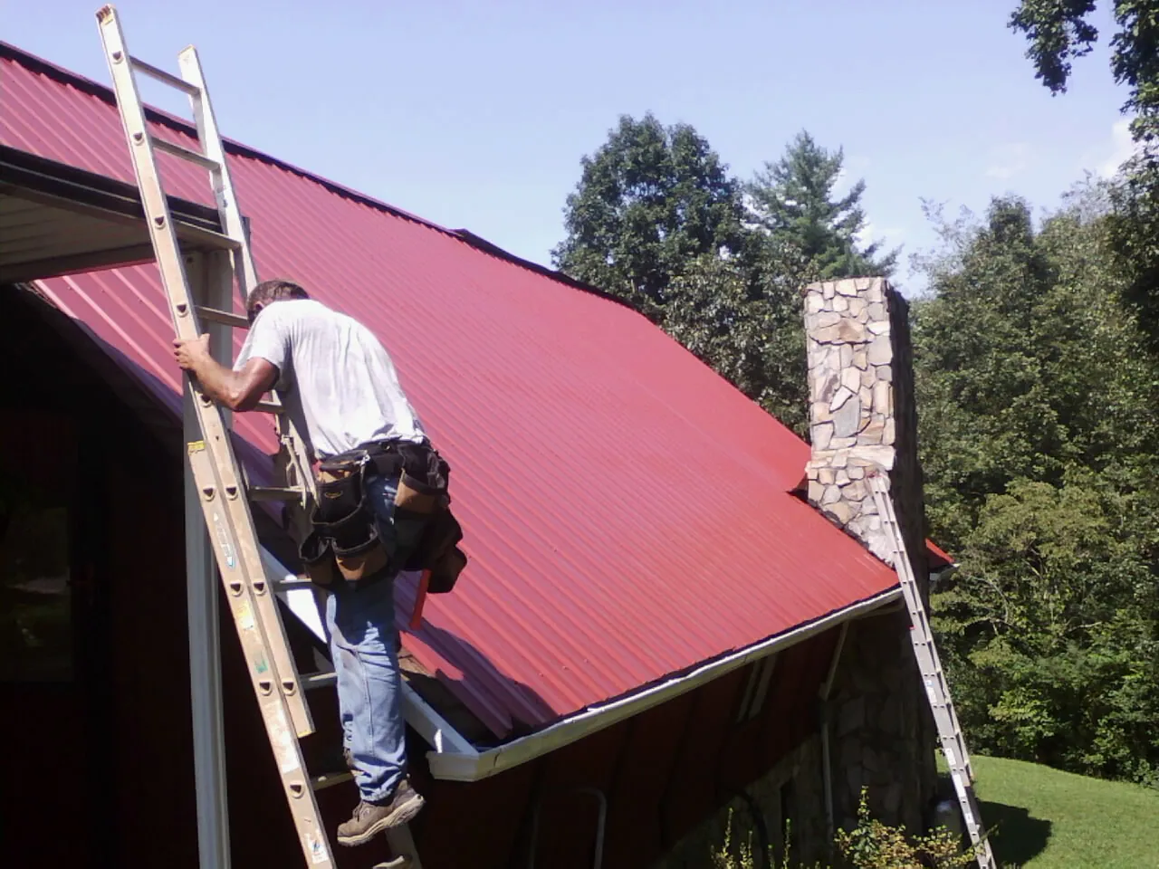 Roofer on a ladder beside a red metal roof and stone chimney — Soffit & Fascia Repair by Roofing Brothers in Renville